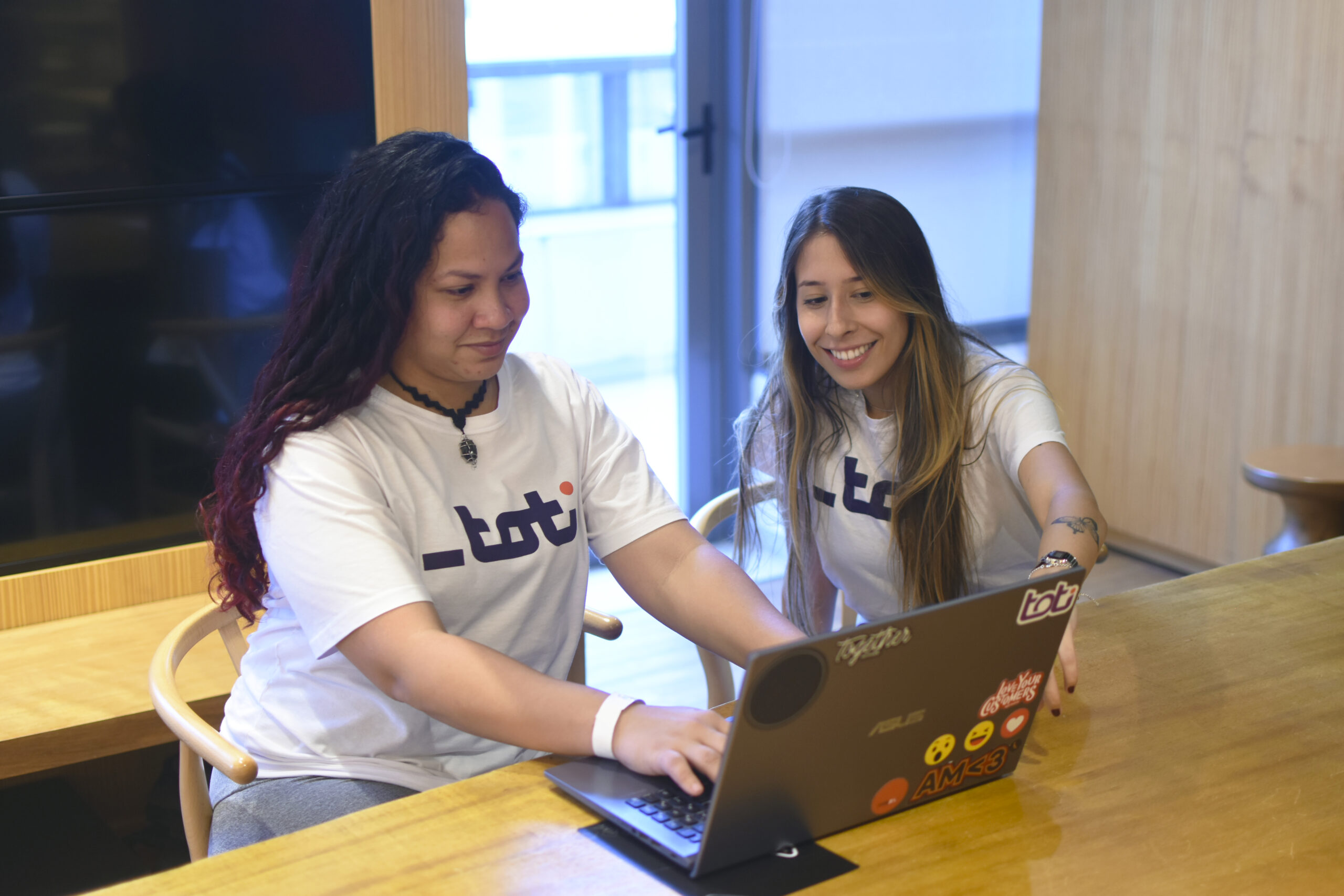 Duas mulheres sorridentes, vestindo camisetas brancas com o logotipo da Toti, trabalham juntas em um laptop. Elas estão sentadas lado a lado em um ambiente interno iluminado.