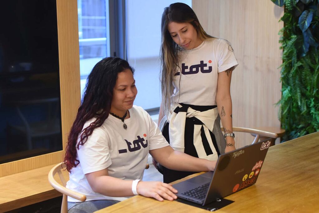 Duas mulheres da Toti colaboram em um laptop em um escritório acolhedor, exemplificando trabalho em equipe e engajamento em um ambiente de escritório moderno.