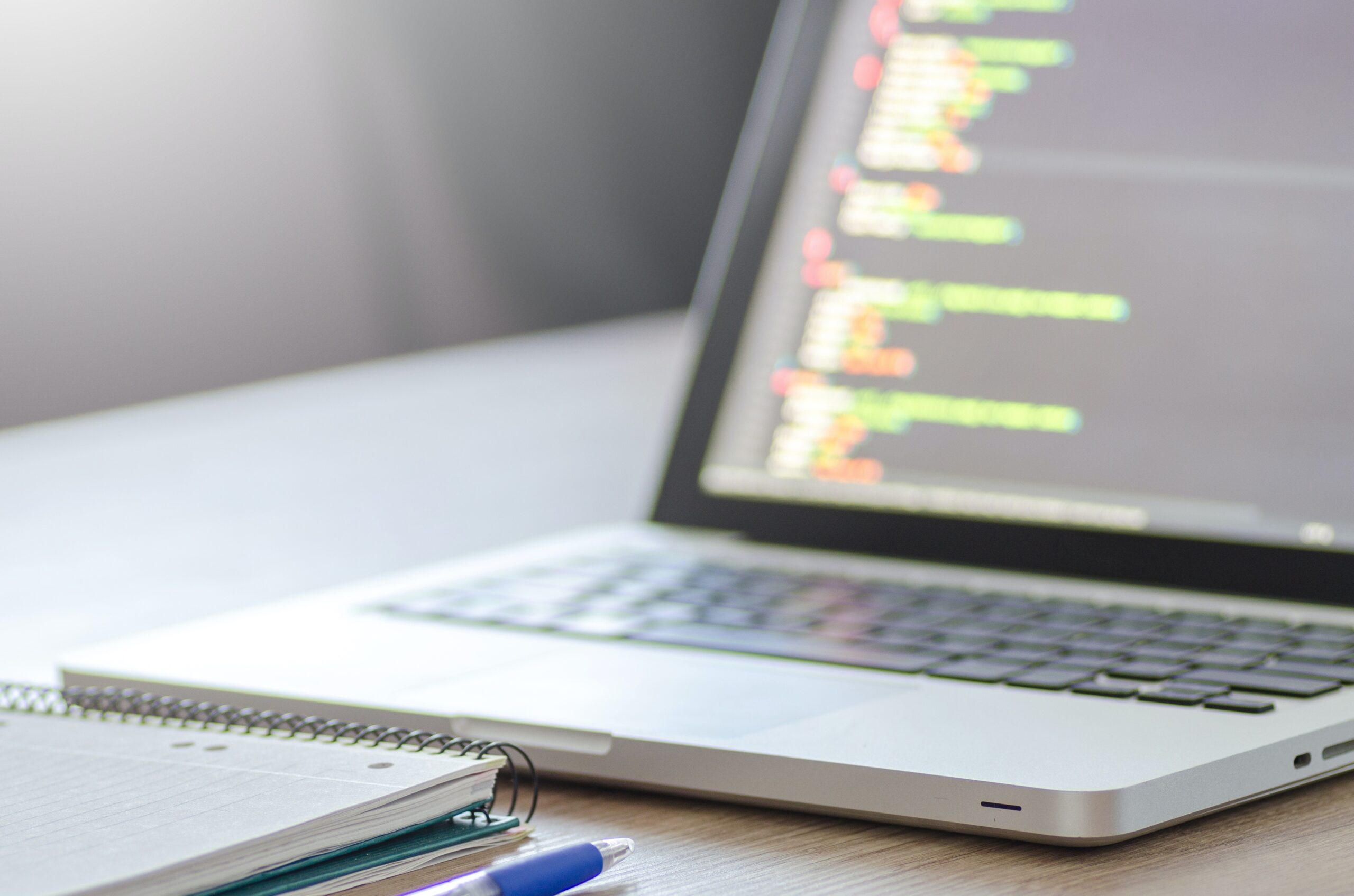 Notebook em uma mesa exibe códigos de programação. Na mesa, também há um caderno e uma caneta.