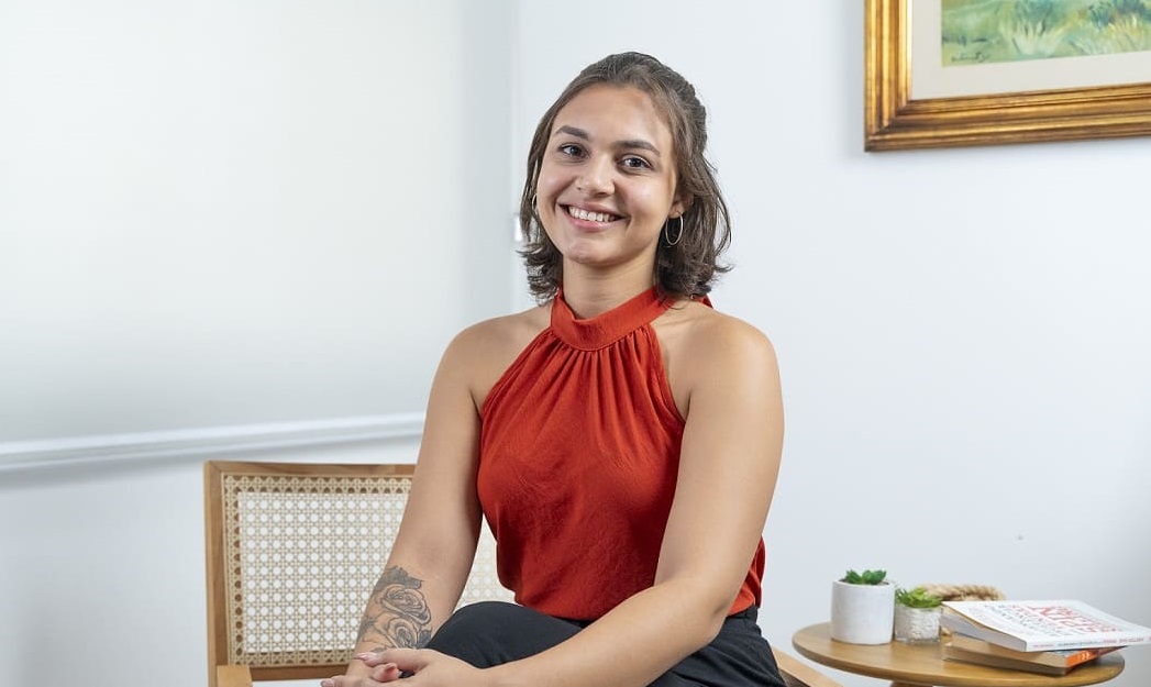 Mulher sentada em uma cadeira sorrindo. Ela é branca, tem cabelo preto curto e está vestindo uma blusa terracota.