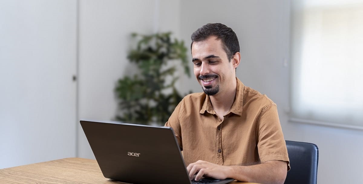 Homem sentado à mesa utilizando um notebook. Ele é branco, tem cabelo curto e preto, e veste uma camisa bege de manga curta.