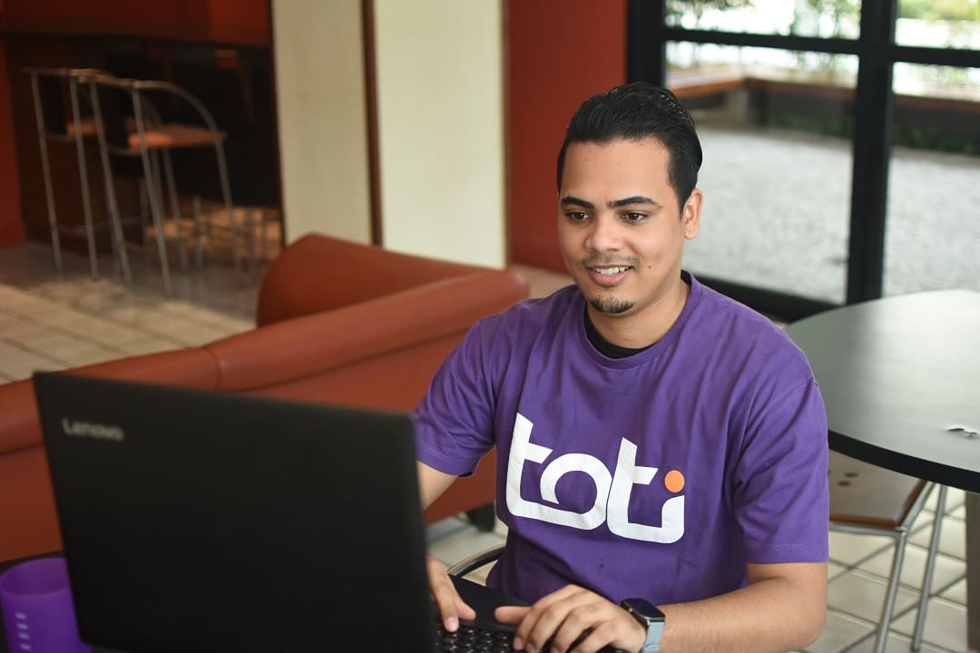 Um homem jovem veste uma camisa roxa com o logotipo da Toti Diversidade. Ele está sentado em uma cadeira, utilizando um notebook.