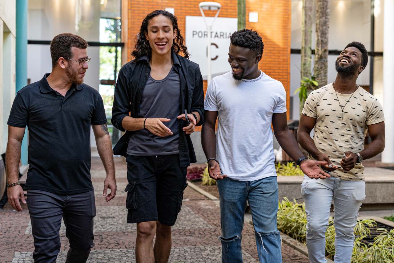 Quatro migrantes do sexo masculino passeiam em um shopping no Brasil. Todos estão andando, conversando e sorrindo.