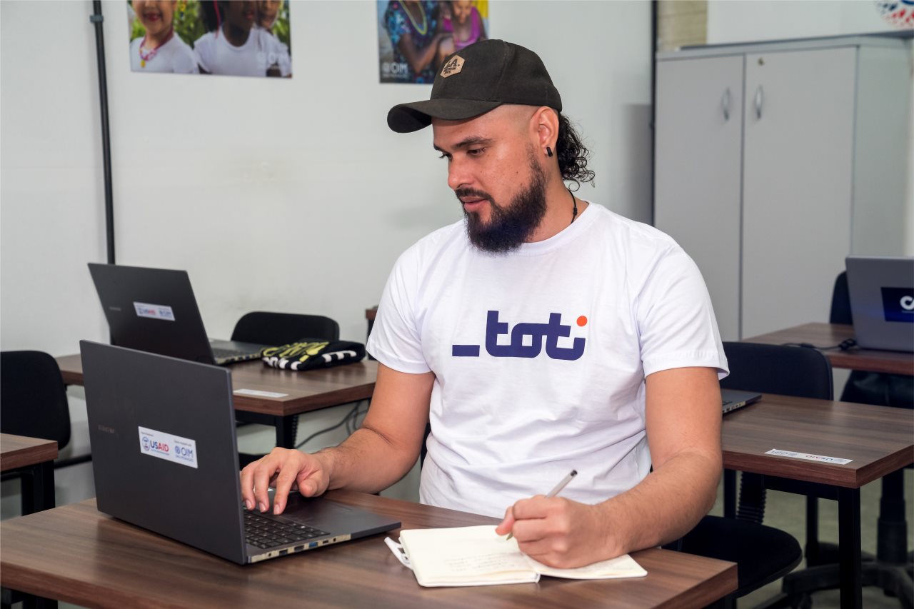 Homem em camiseta da Toti e boné está concentrado em seu laptop enquanto toma notas em um caderno, em uma sala de aula, indicando um ambiente de aprendizado.