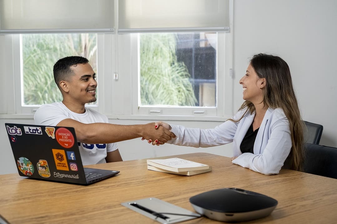 Duas pessoas apertam as mãos em um gesto de concordância mútua. Uma delas é do sexo masculino e a outra é do sexo feminino. Ambos estão em um ambiente corporativo. Na mesa em que estão sentados, há um notebook.