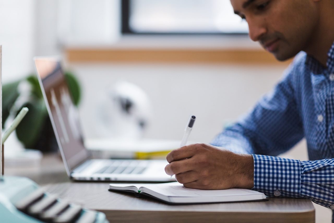 Homem em camisa xadrez azul concentrado escrevendo em um caderno em sua mesa de trabalho, com um laptop aberto à sua frente, em um ambiente de escritório focado e produtivo.