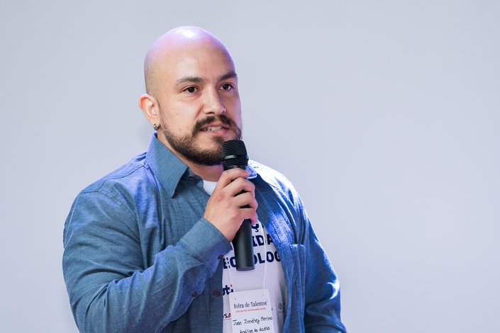 Um homem, vestindo camisa jeans azul, fala em um microfone durante um evento, destacando-se contra um fundo neutro e iluminado.