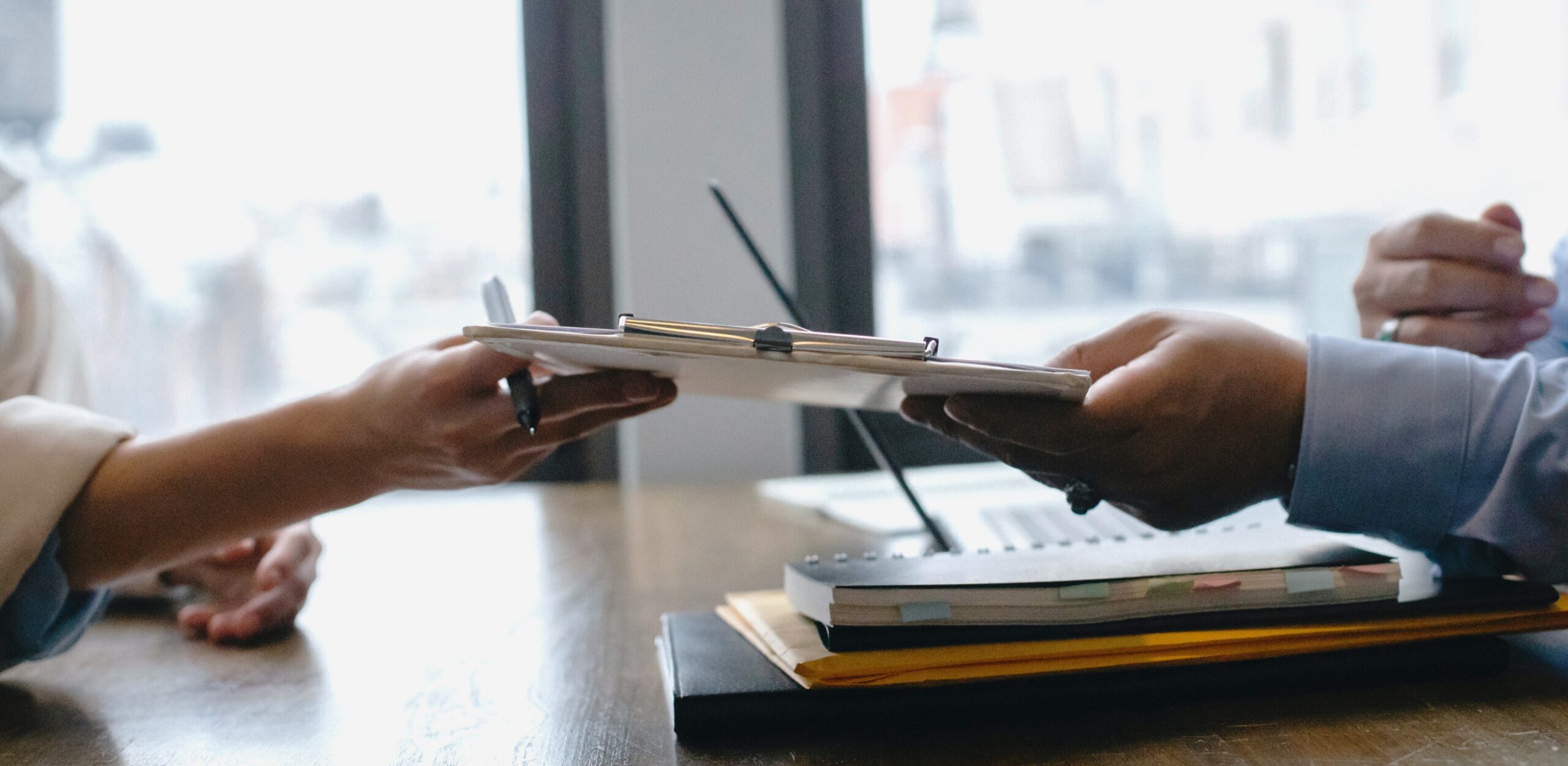 Duas pessoas seguram uma prancheta com algum documento. Na foto, apenas aparecem as mãos dessas pessoas. Na mesa em que elas estão sentadas, aparecem alguns documentos. É provável que estejam em uma entrevista de emprego falando sobre pretensão salarial.