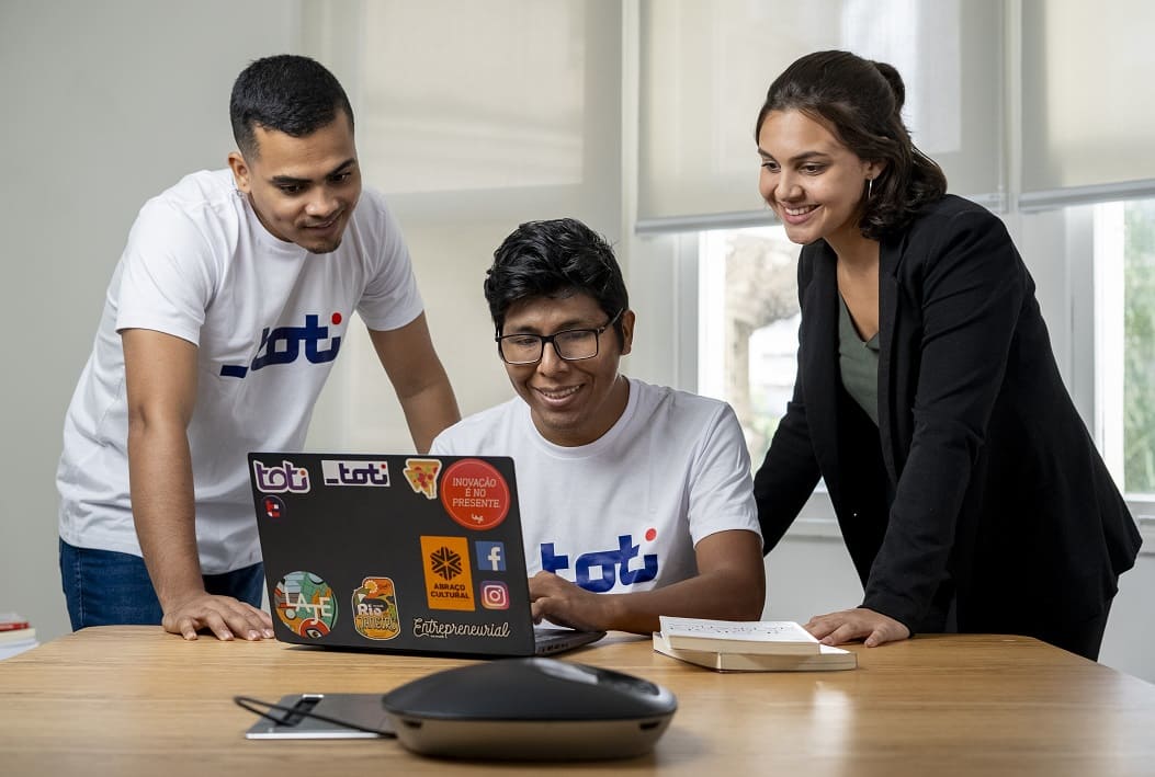 Três jovens, sendo dois homens e uma mulher, sorriem enquanto olham para um notebook. Um dos homens está sentado em uma cadeira e mexendo no notebook.