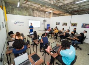 Um ambiente de sala de aula com um professor ao lado de um projetor. Os alunos estão sentados em carteiras e usando computadores para acompanhar a aula.