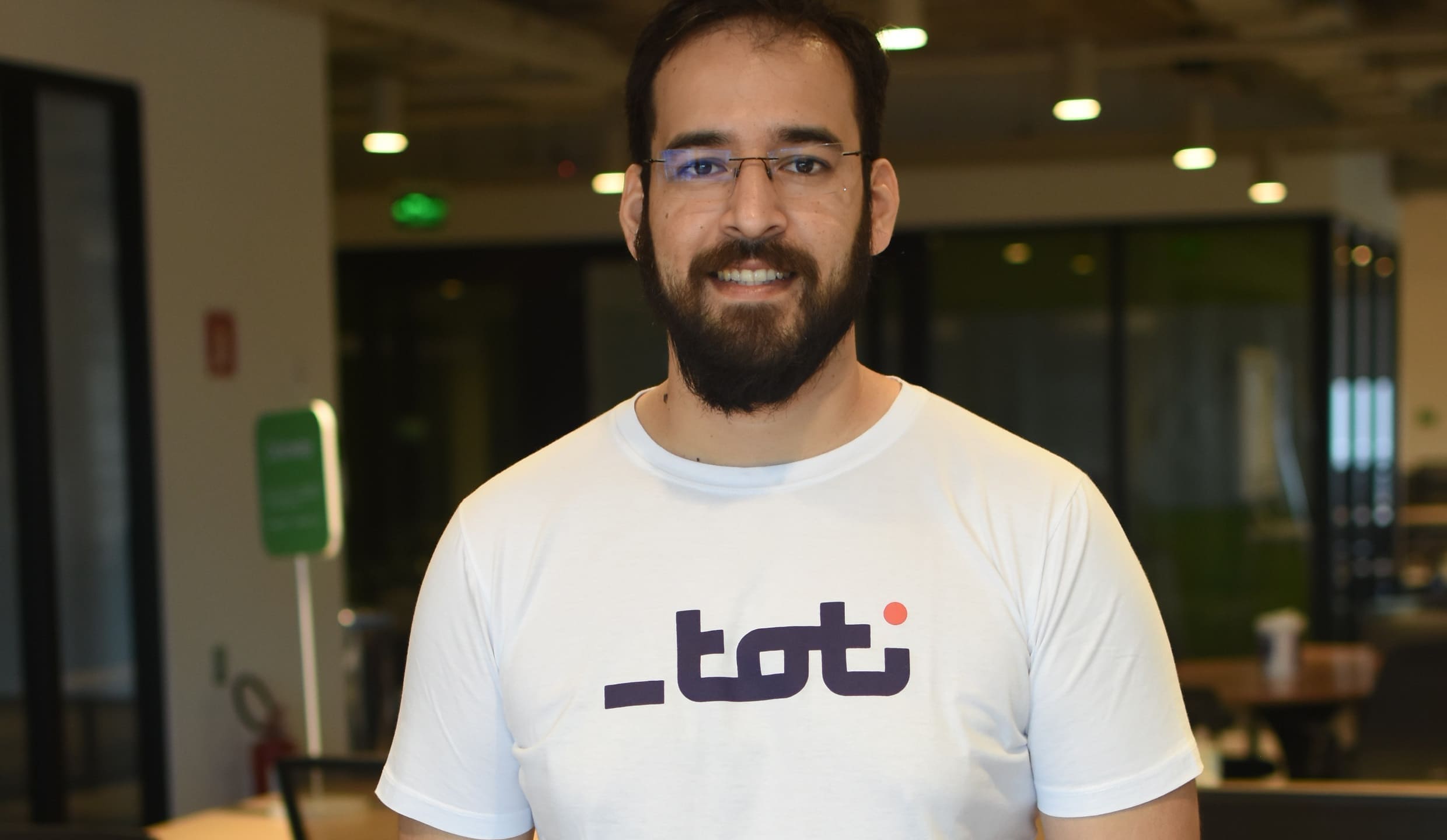 Homem sorridente com camiseta da Toti posa em um escritório moderno, exibindo um ambiente de trabalho casual e acolhedor.