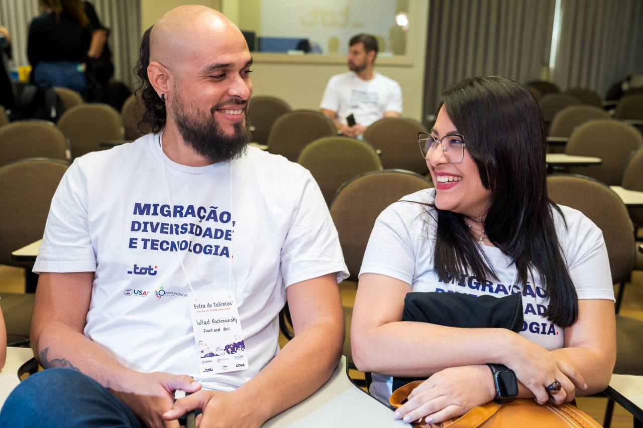 Na foto, duas pessoas estão conversando e sorrindo. Uma delas é Carmen, uma mulher parda de cabelos pretos, natural da Venezuela. A outra pessoa é Wladimir, um homem venezuelano com barba. Ambos estavam em um evento para procurar trabalho para imigrantes no Brasil.