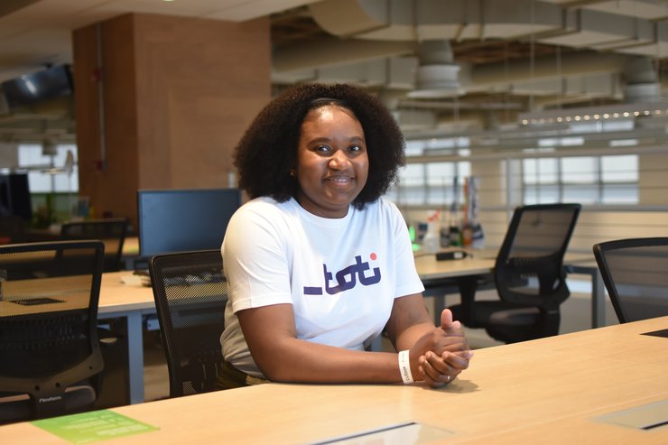 Bianda é uma pessoa do sexo feminino, negra e do Haiti. Na imagem, ela está sentada em uma mesa dentro de um ambiente corporativo. Ela veste uma camisa com a logo da Toti e sorri para a câmera.