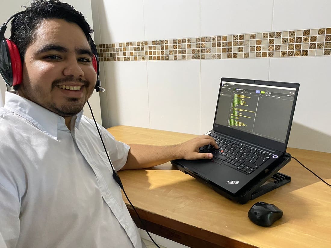 Na foto, aparece Wendell Rojas, um migrante que fez uma transição de carreira no Brasil. Wendell é branco e venezuelano. Ele está sentado usando um notebook, que exibe um código de programação na tela.