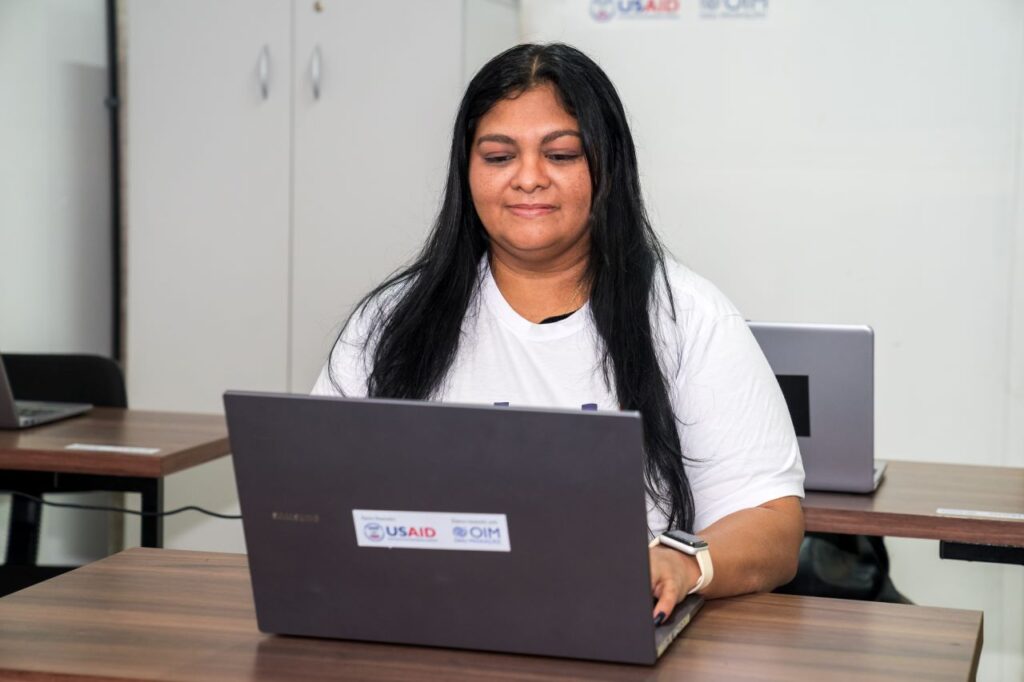 Mulher usando um laptop em um ambiente de treinamento, com adesivos da USAID e OIM no laptop, sugerindo envolvimento em programas para imigrantes ou refugiados, em um cenário de aprendizado ou capacitação.