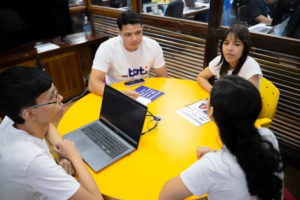 Quatro pessoas estão reunidas em torno de uma mesa redonda amarela, em um ambiente de escritório. Elas estão vestindo camisetas brancas com o logotipo da Toti. Há um notebook sobre a mesa, além de materiais impressos com informações e gráficos, sugerindo uma discussão de trabalho.