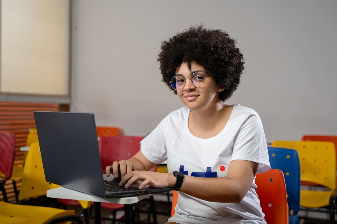 Mulher jovem em uma sala de aula, utilizando um notebook e vestindo uma camisa da Toti, representando foco no aprendizado e na formação profissional.