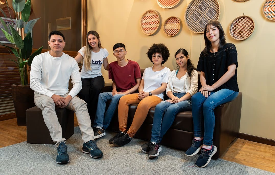 Grupo de jovens sentados em um ambiente acolhedor, representando diversidade cultural. Ao fundo, uma parede decorada com cestos artesanais, transmitindo elementos de tradição e inclusão.