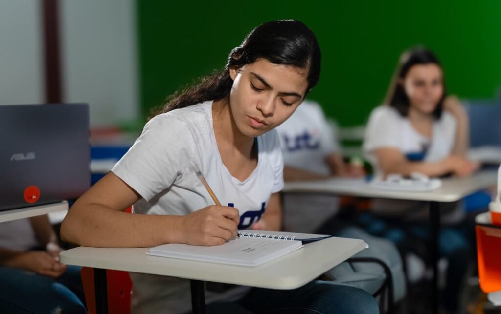 Mulher jovem concentrada escrevendo em um caderno durante uma aula. Ao fundo, outras pessoas também participam da atividade em um ambiente educacional.