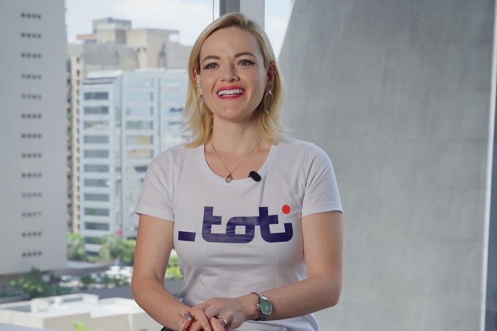 Mulher sorrindo, vestindo uma camiseta branca com o logo da Toti, sentada em um ambiente moderno com janelas amplas e vista urbana ao fundo.