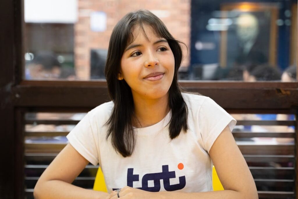 Jovem sorridente usando uma camiseta com o logo da Toti, sentada em uma mesa amarela em um ambiente descontraído com fundo desfocado.