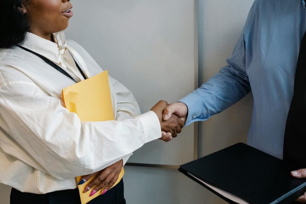 Duas pessoas apertando as mãos em um ambiente profissional. Uma delas segura uma pasta amarela e a outra, um portfólio preto, sugerindo o contexto de uma entrevista de emprego ou reunião de negócios.