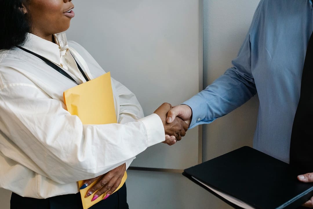 Duas pessoas apertando as mãos em um ambiente profissional. Uma delas segura uma pasta amarela e a outra, um portfólio preto, sugerindo o contexto de uma entrevista de emprego ou reunião de negócios.